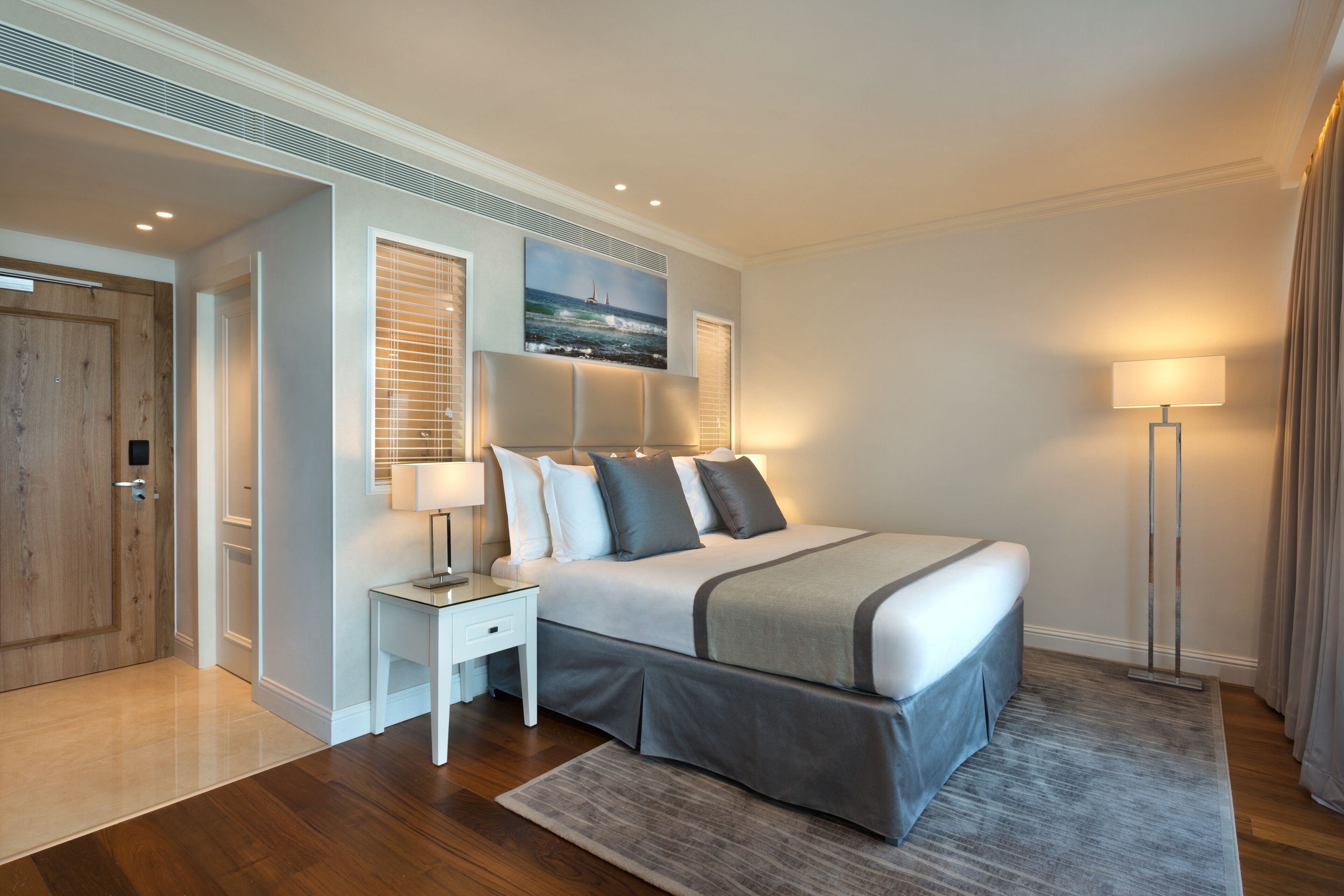 A modern hotel room with a large bed, a bedside table, a lamp, and a painting on the wall.