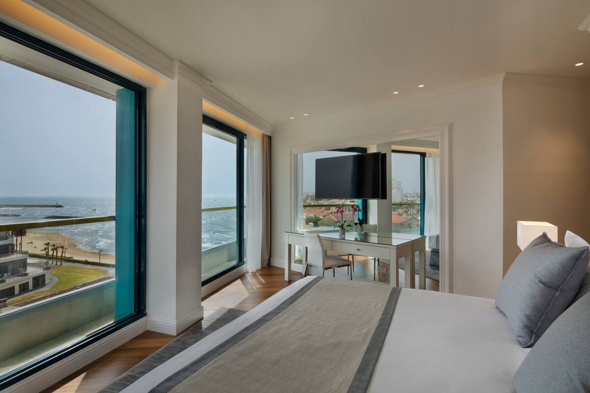A modern hotel room with a large bed, a desk with a TV, and a view of the ocean.