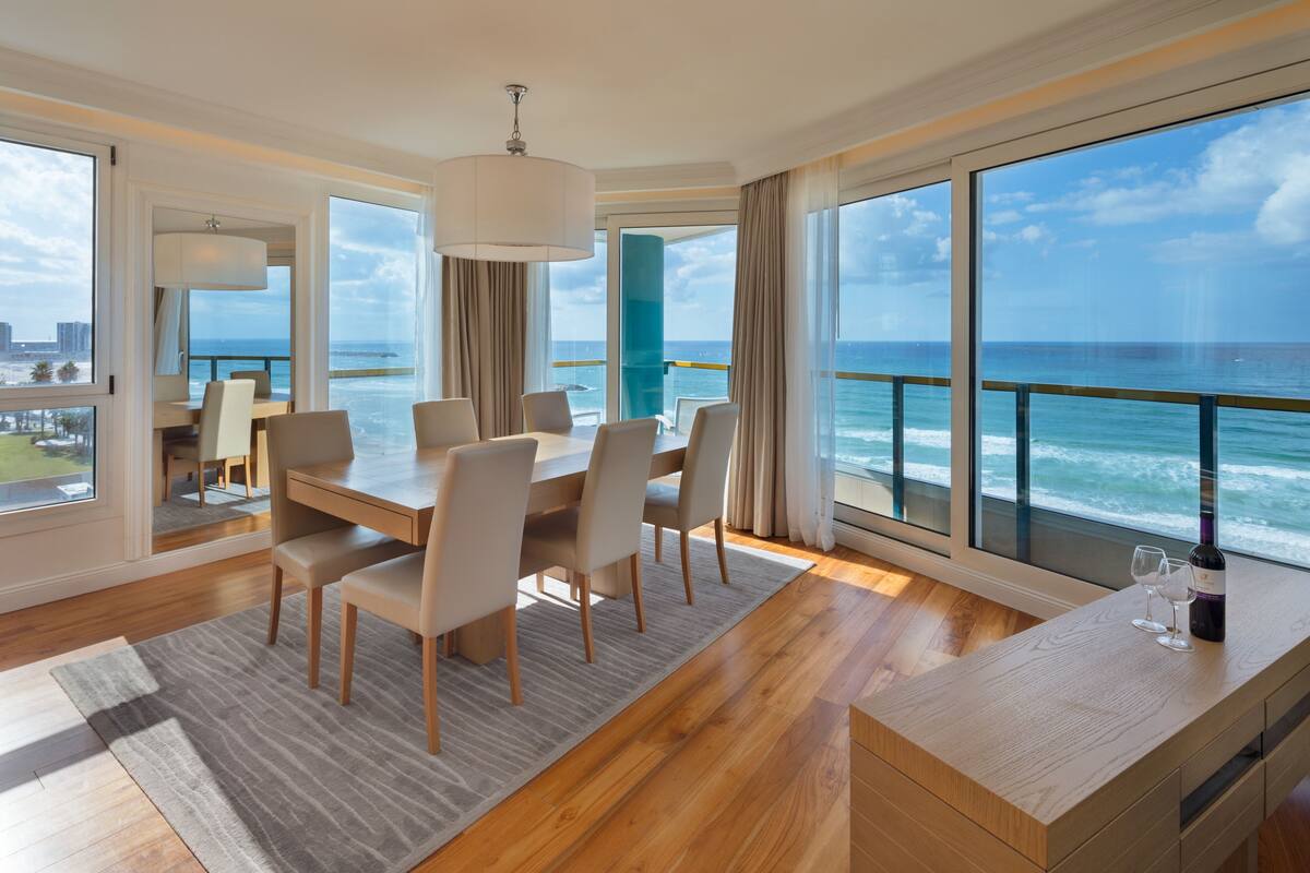 A modern hotel room with a large ocean view, a dining area with a wooden table and chairs, and a minibar with wine glasses.