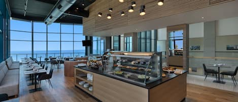 A modern restaurant with a buffet, wooden flooring, and large windows offering a view of the ocean.