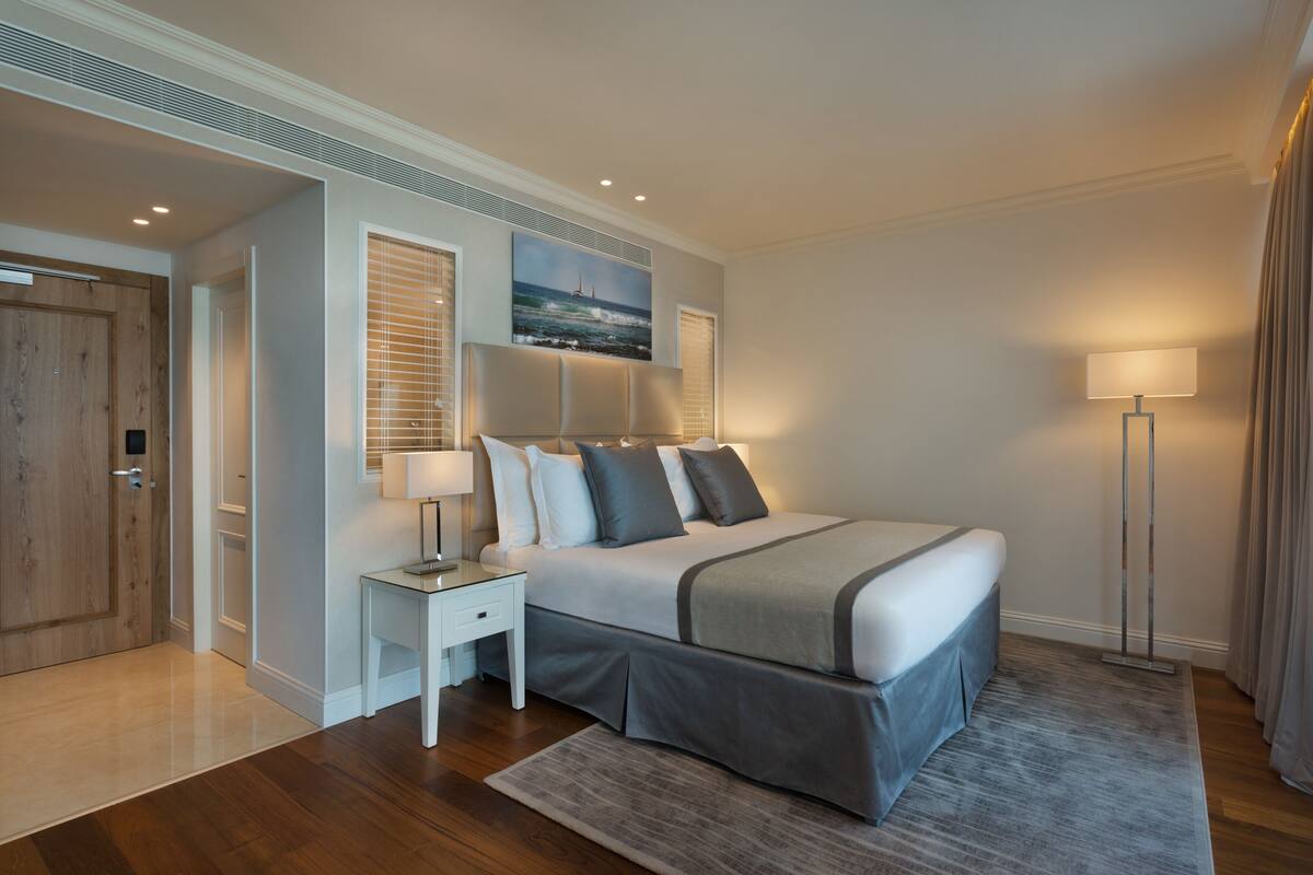 A modern bedroom with a large bed, bedside tables, a nightstand, a painting on the wall, and a floor lamp.