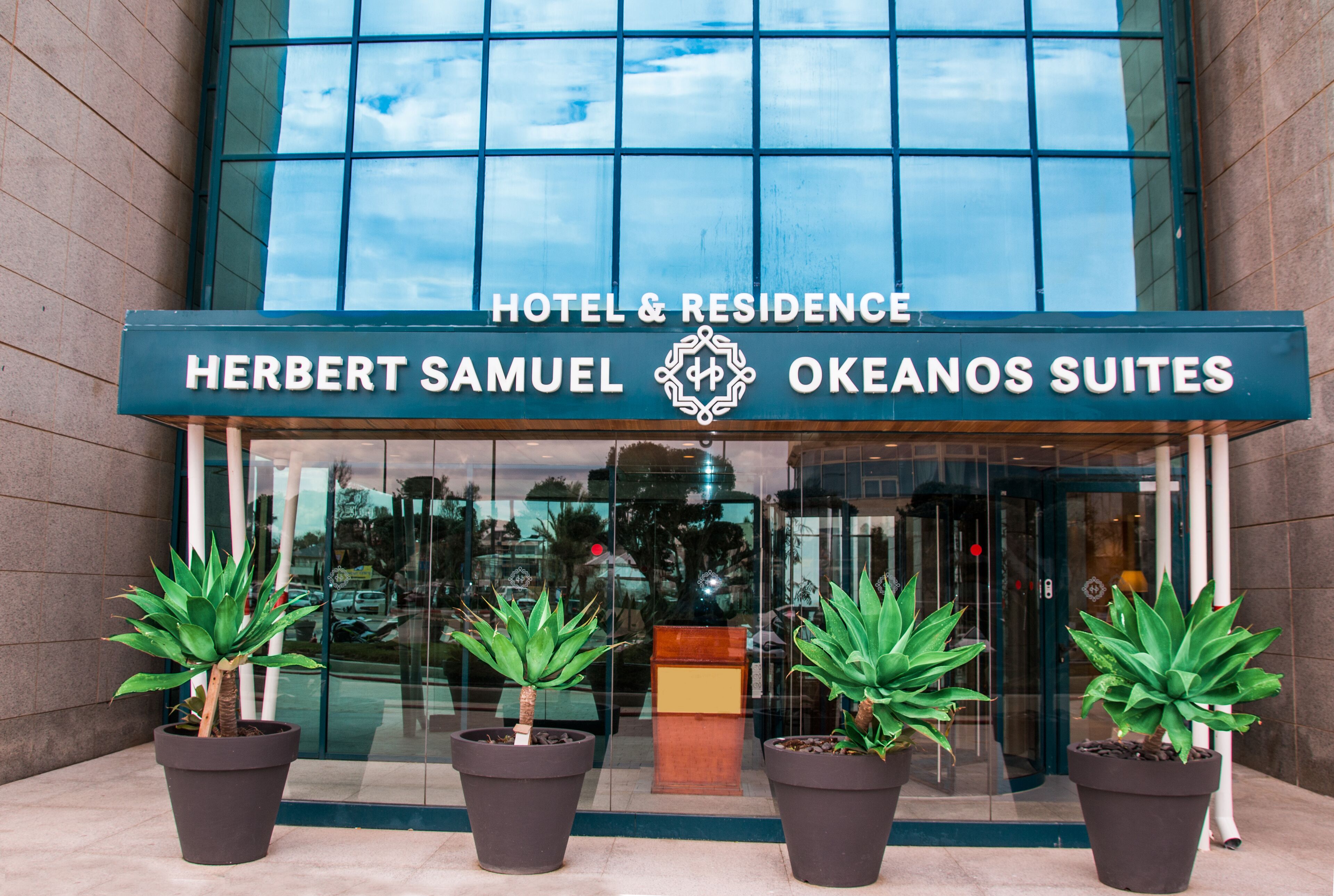 The entrance of Herbert Samuel Okeanos Suites, featuring a modern glass facade and four potted plants in front.