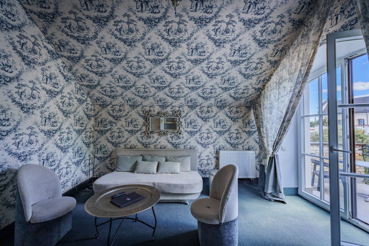 Een kamer met behang met een patroon, een bank, twee fauteuils, een kleine ronde tafel en een groot raam met uitzicht.