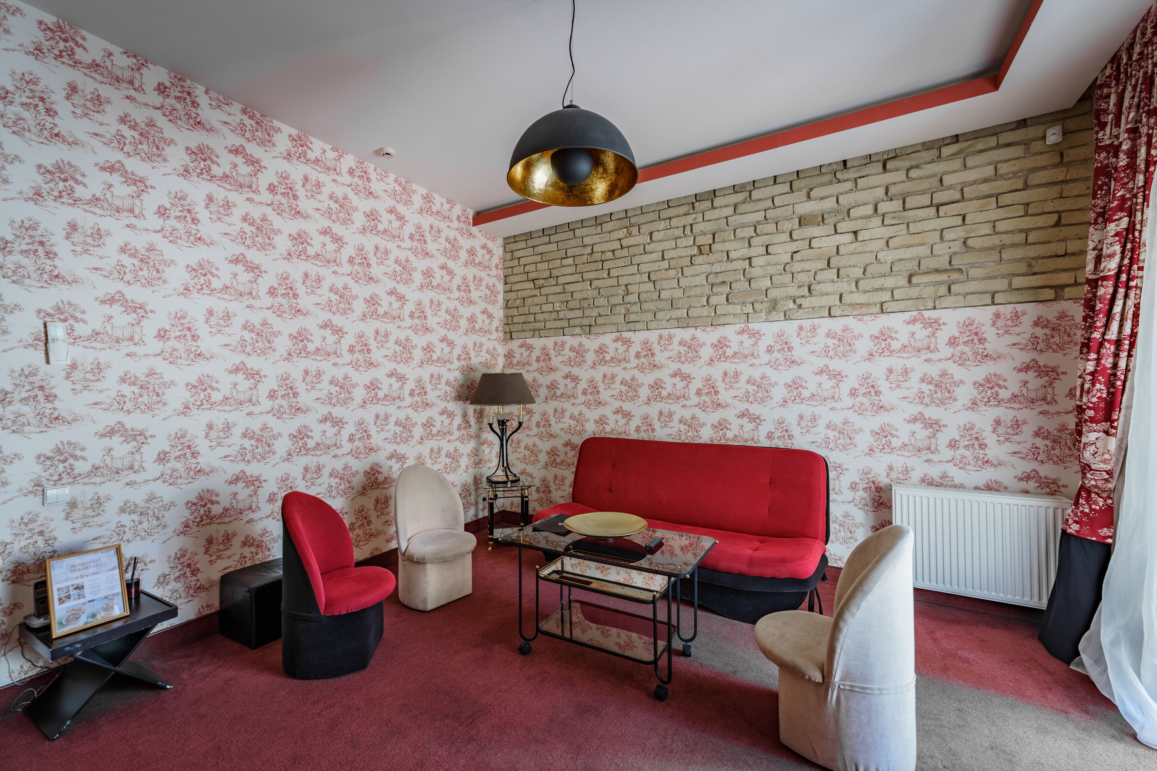 A room with a red sofa, two armchairs, a small table, a lamp, and a brick wall.