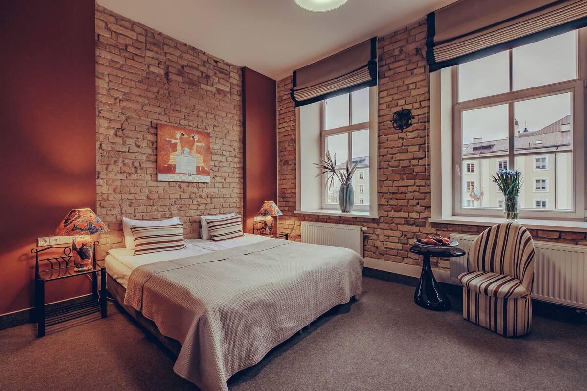 Slaapkamer met een bakstenen muur, een groot raam, een bed met een gestreept kussen, een stoel en een klein tafeltje met een lamp.