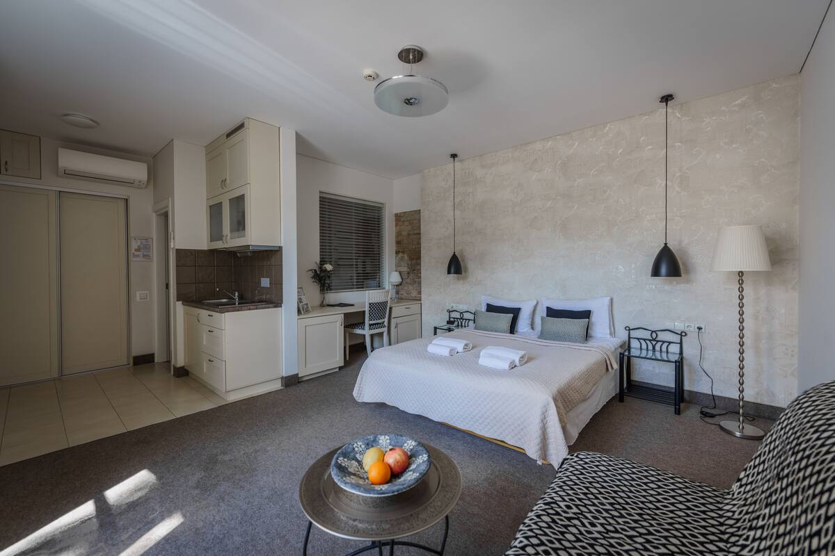 Een moderne hotelkamer met kitchenette, een bed met wit dekbedovertrek, nachtkastje met fruitschaal en een gestreept bankje.