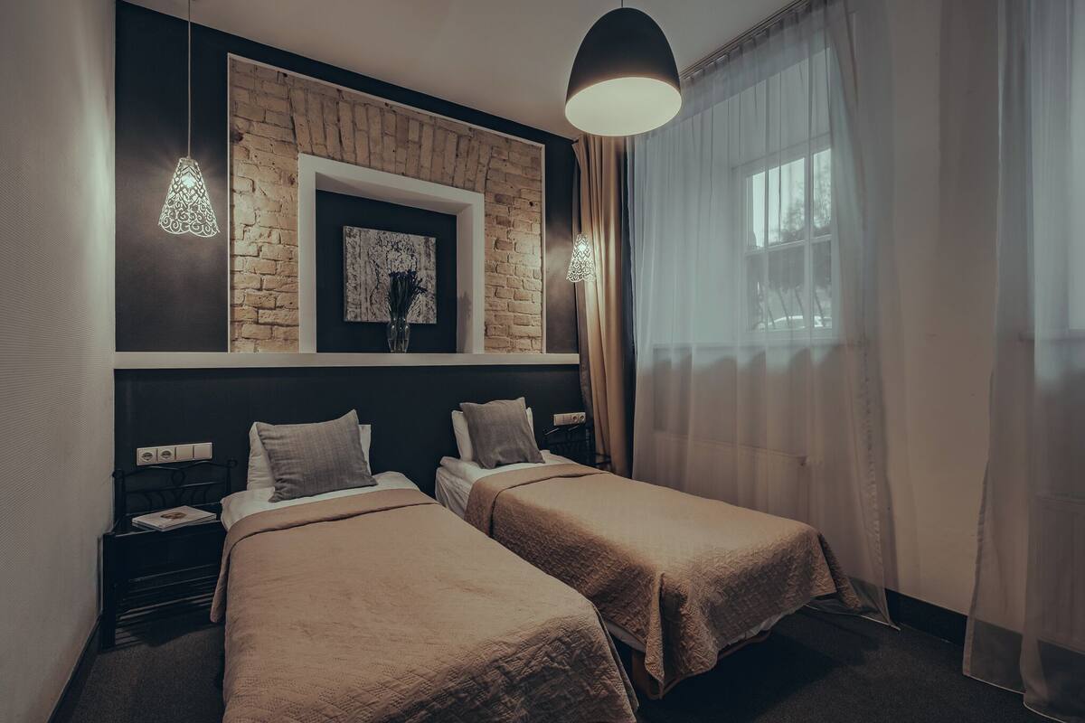 Een hotelkamer met twee bedden, een bakstenen muur, een ingelijste afbeelding en een groot raam met dunne gordijnen.