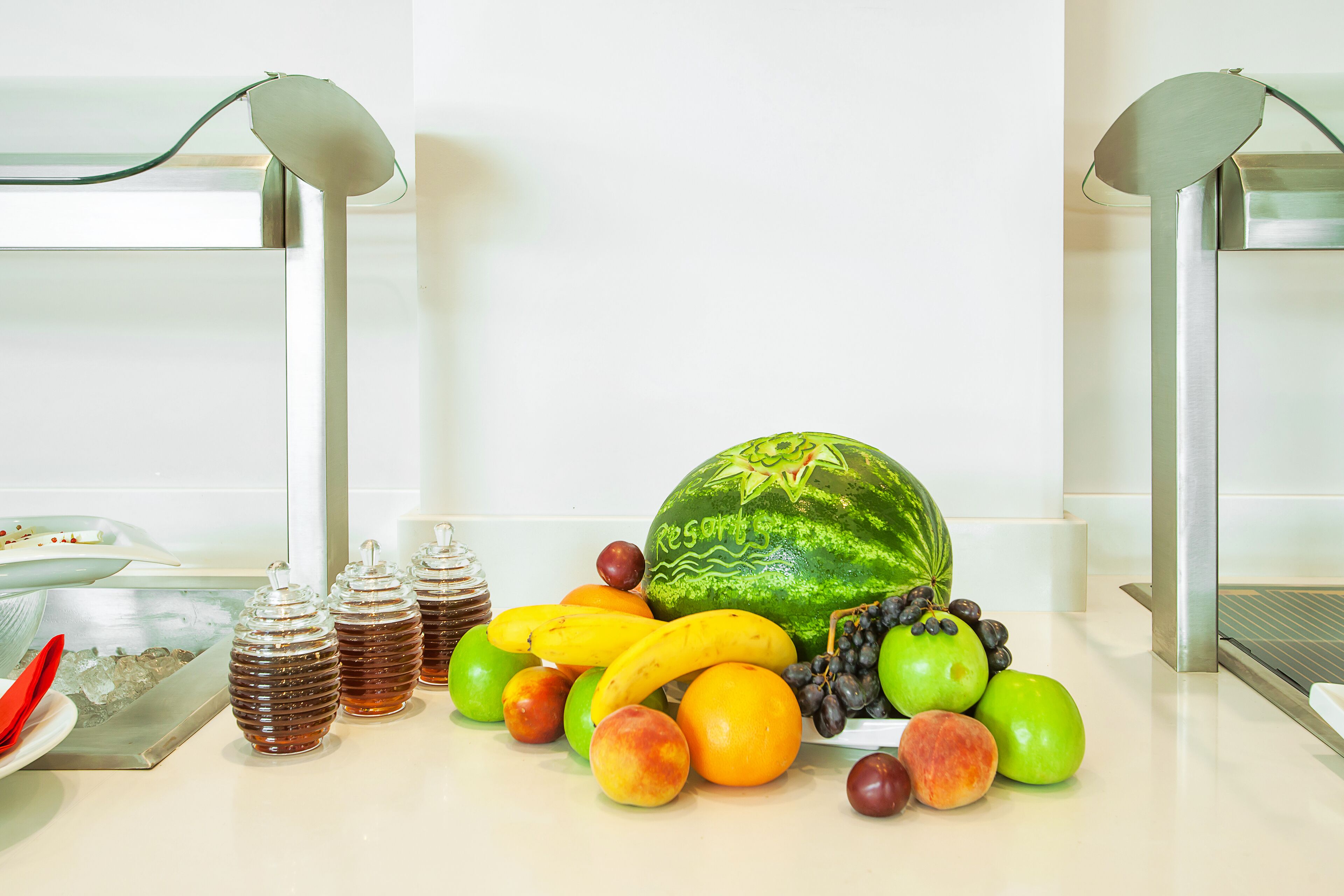Una exhibición de frutas con sandía, plátanos, manzanas y uvas sobre una barra.