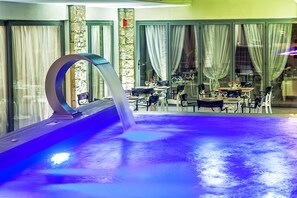 An indoor swimming pool with a modern design, surrounded by glass walls and curtains, and furnished with tables and chairs.