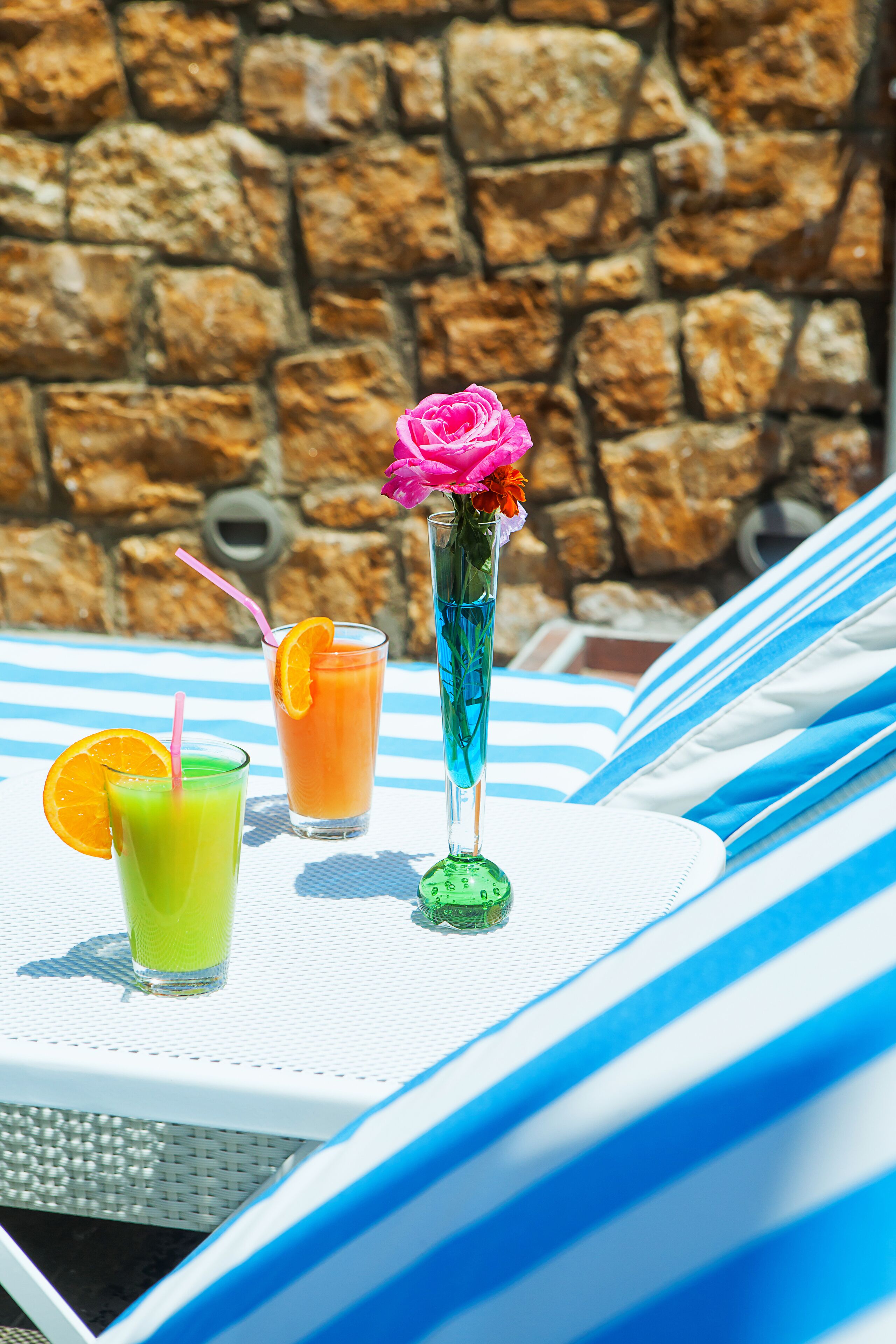 A table with three drinks, a flower, and a striped lounge chair.