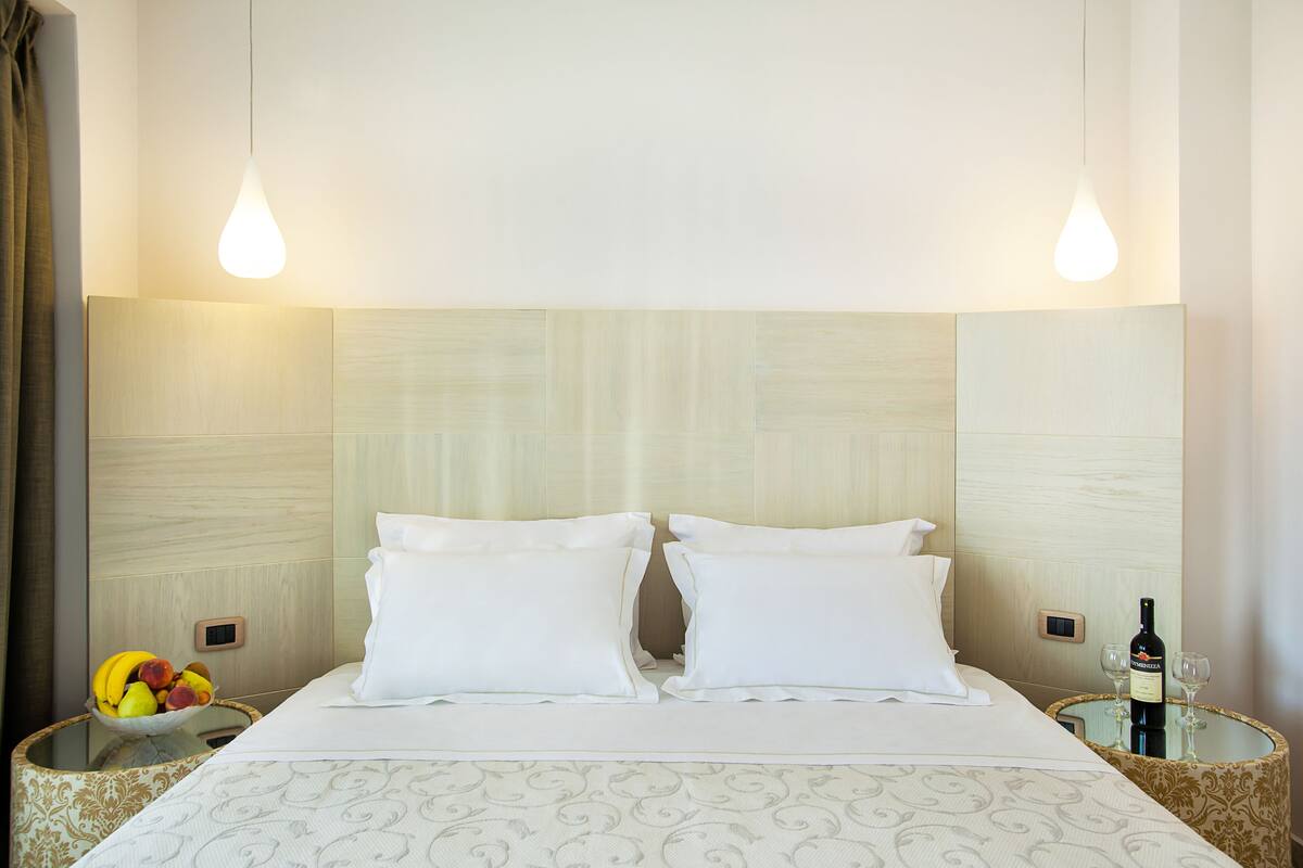 A hotel room with a bed, two bedside tables, a fruit bowl, and a wine bottle.