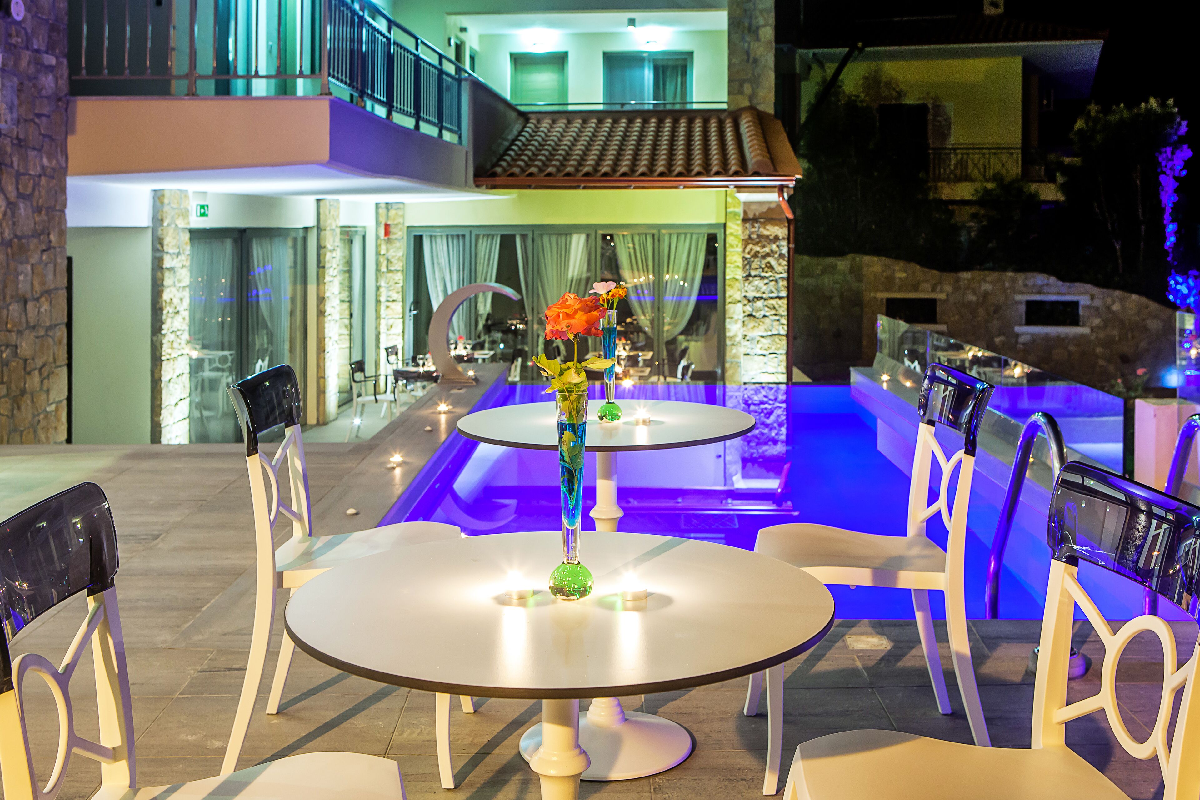 An outdoor dining area with round tables and chairs, a decorative vase with flowers, and a pool with blue lighting in the background.
