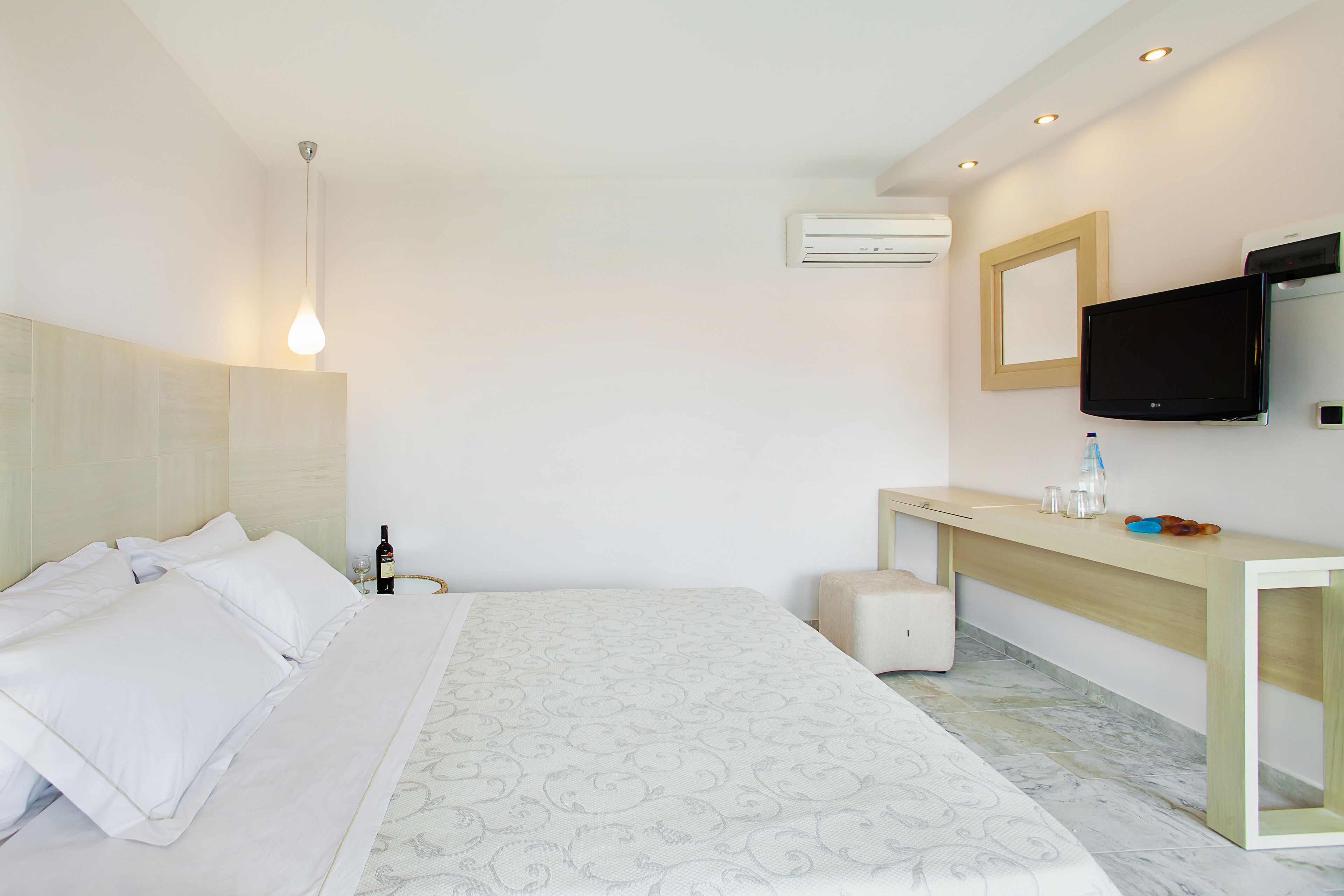 A hotel room with a bed, bedside table, lamp, air conditioning unit, television, and a small stool.