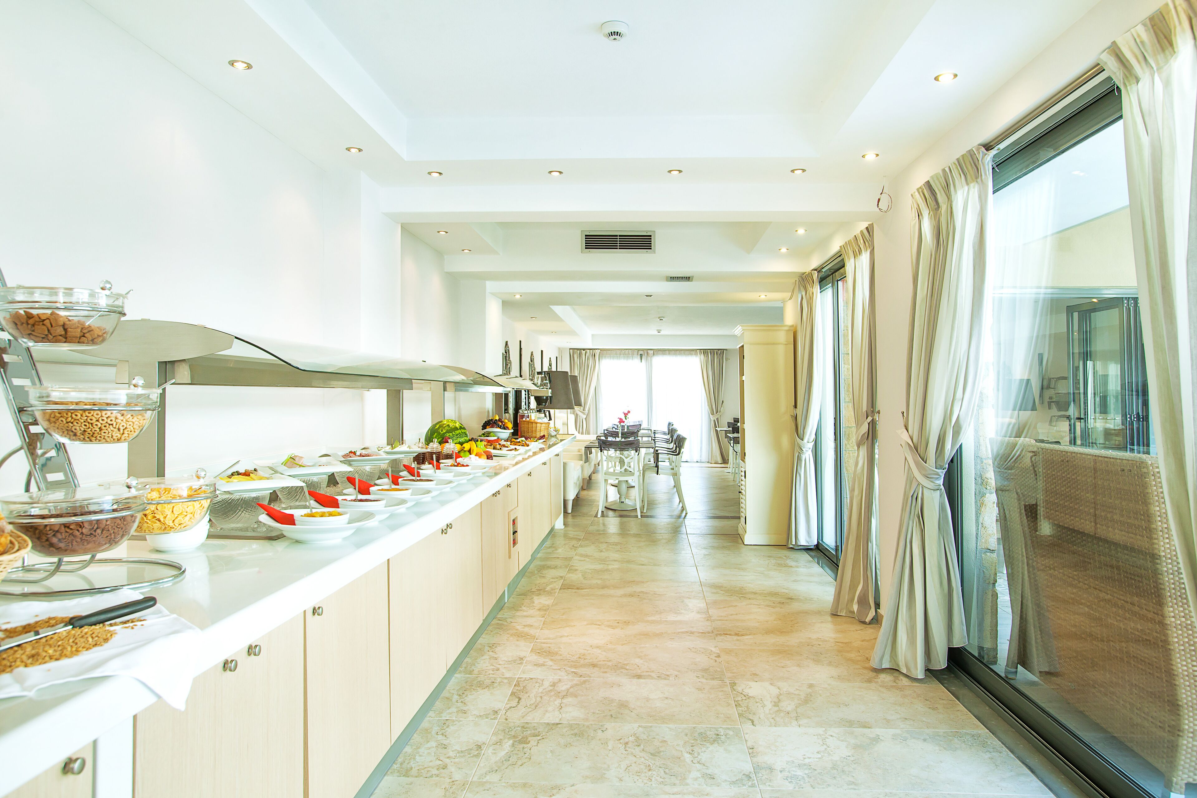 A long, narrow kitchen with a marble floor, a buffet line with various food items, and a view of the dining area through large windows.