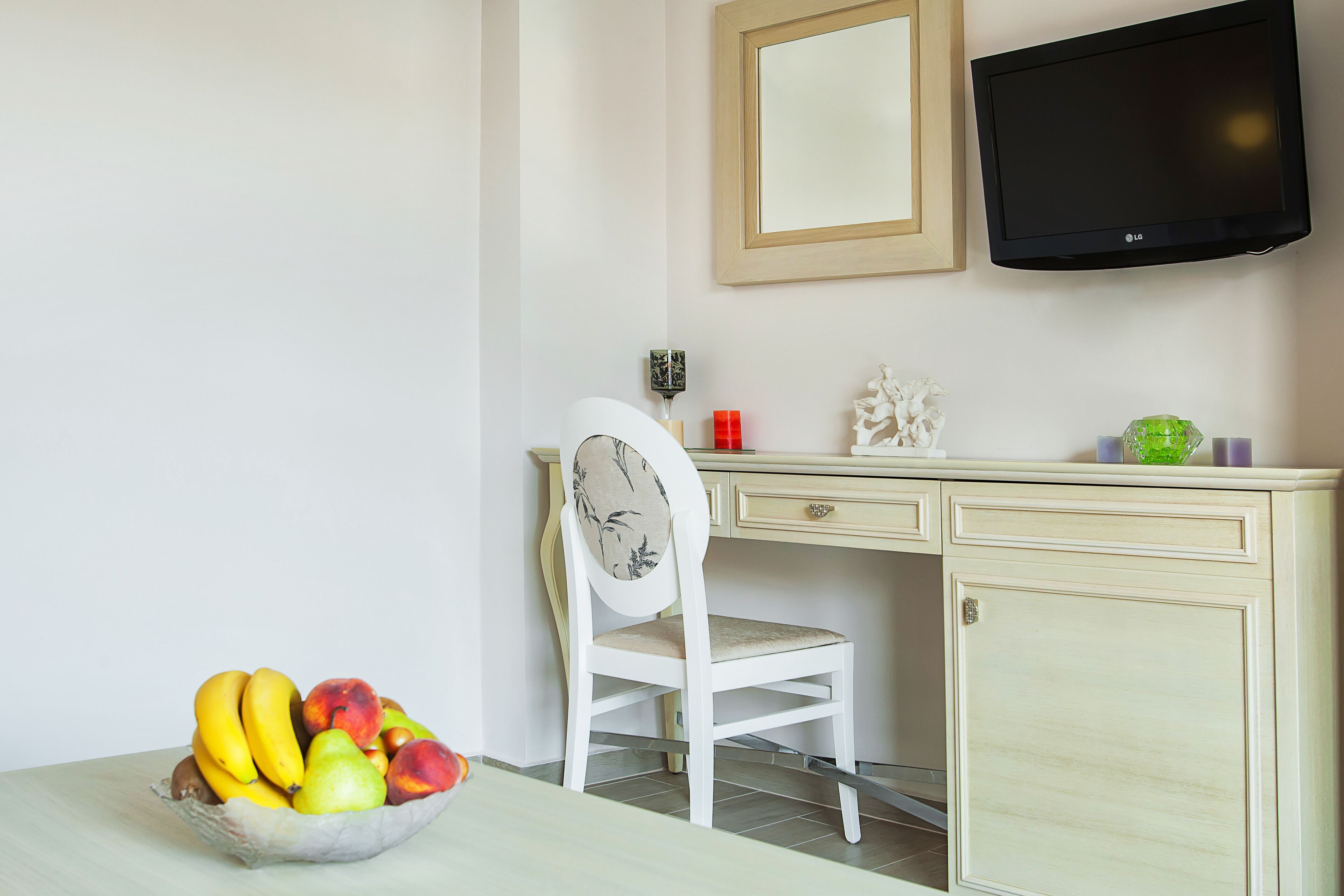 A room with a dining table, a white chair, a fruit bowl, a desk with a computer monitor, and a cabinet.