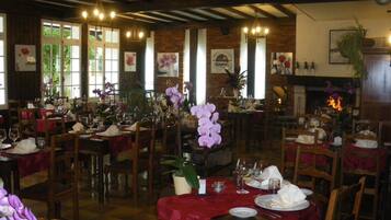 Restaurante com várias mesas preparadas para refeições, apresentando uma parede de tijolos, vigas de madeira e janelas amplas.