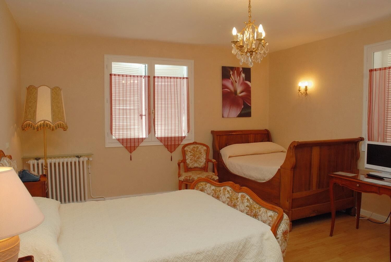 A bedroom with a bed, a wooden chair, a desk with a computer, a radiator, and a chandelier.