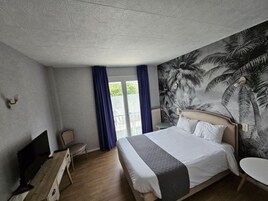 A hotel room with a large bed, a flat-screen TV, a desk, and a chair. There is a balcony with a view of greenery.