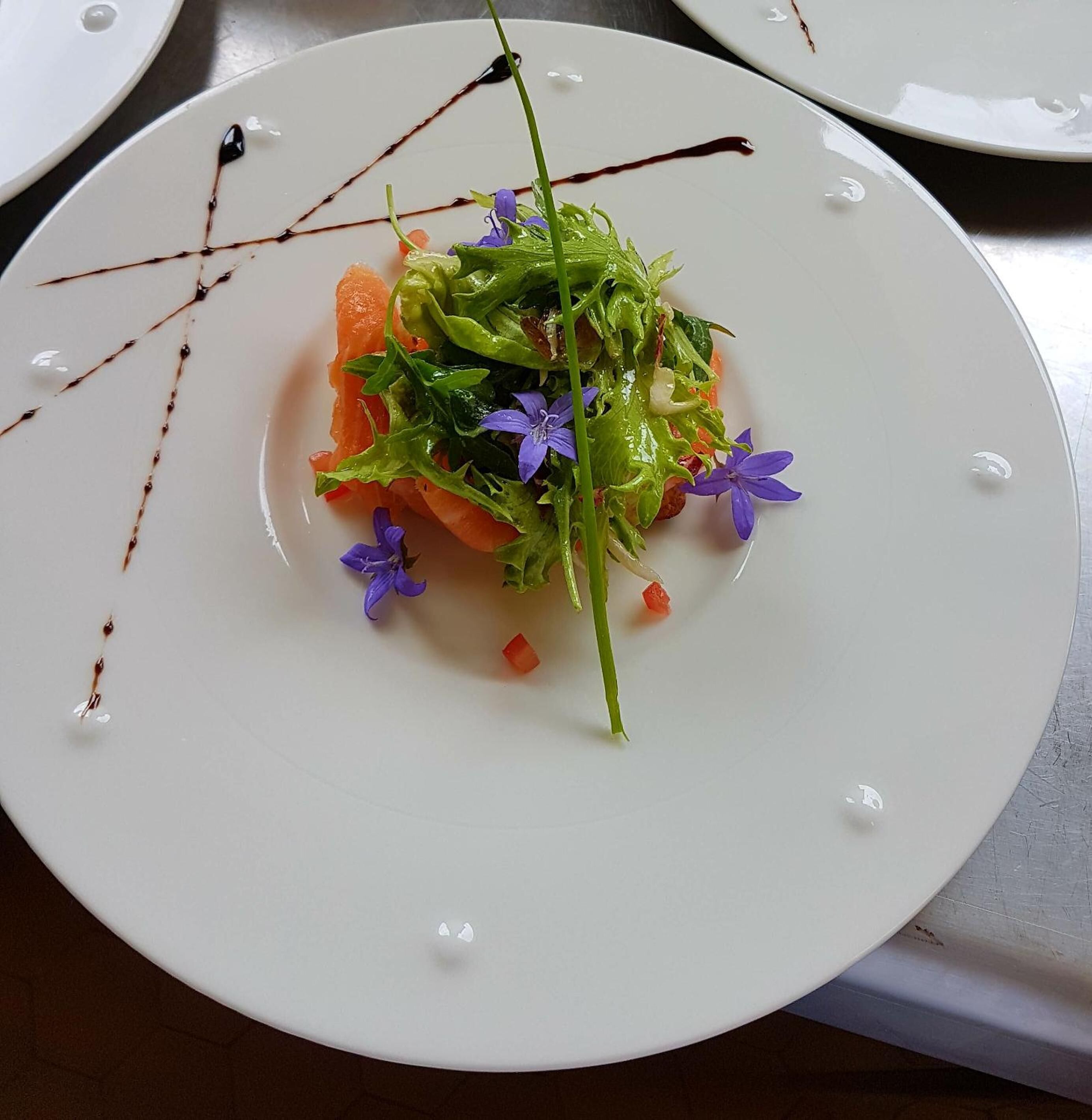 Un plato blanco con ensalada, incluyendo hojas verdes, zanahorias y flores moradas, sobre una mesa.