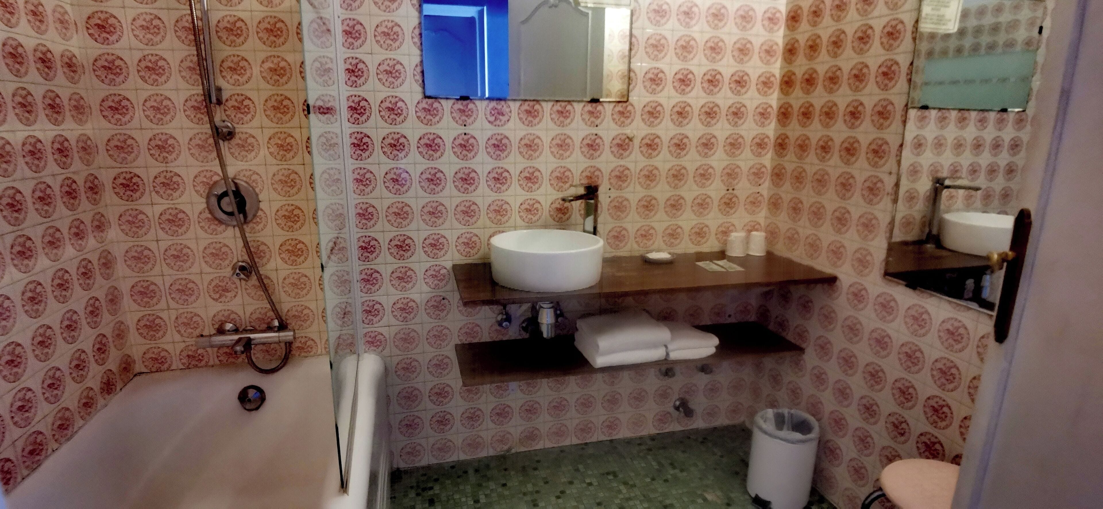 Een badkamer met behang met een patroon, een witte wastafel, een spiegel, een handdoekhouder en een prullenbak.