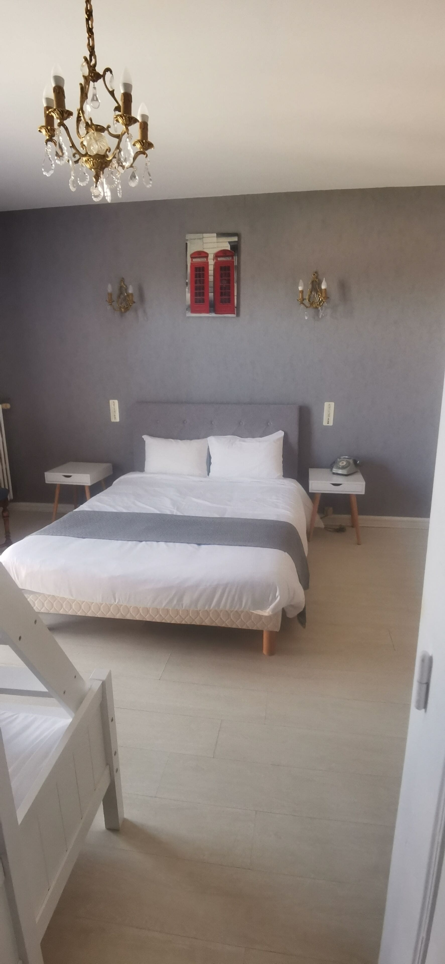 Een slaapkamer met een groot bed, nachtkastjes, een kroonluchter en een rode telefooncel aan de muur.