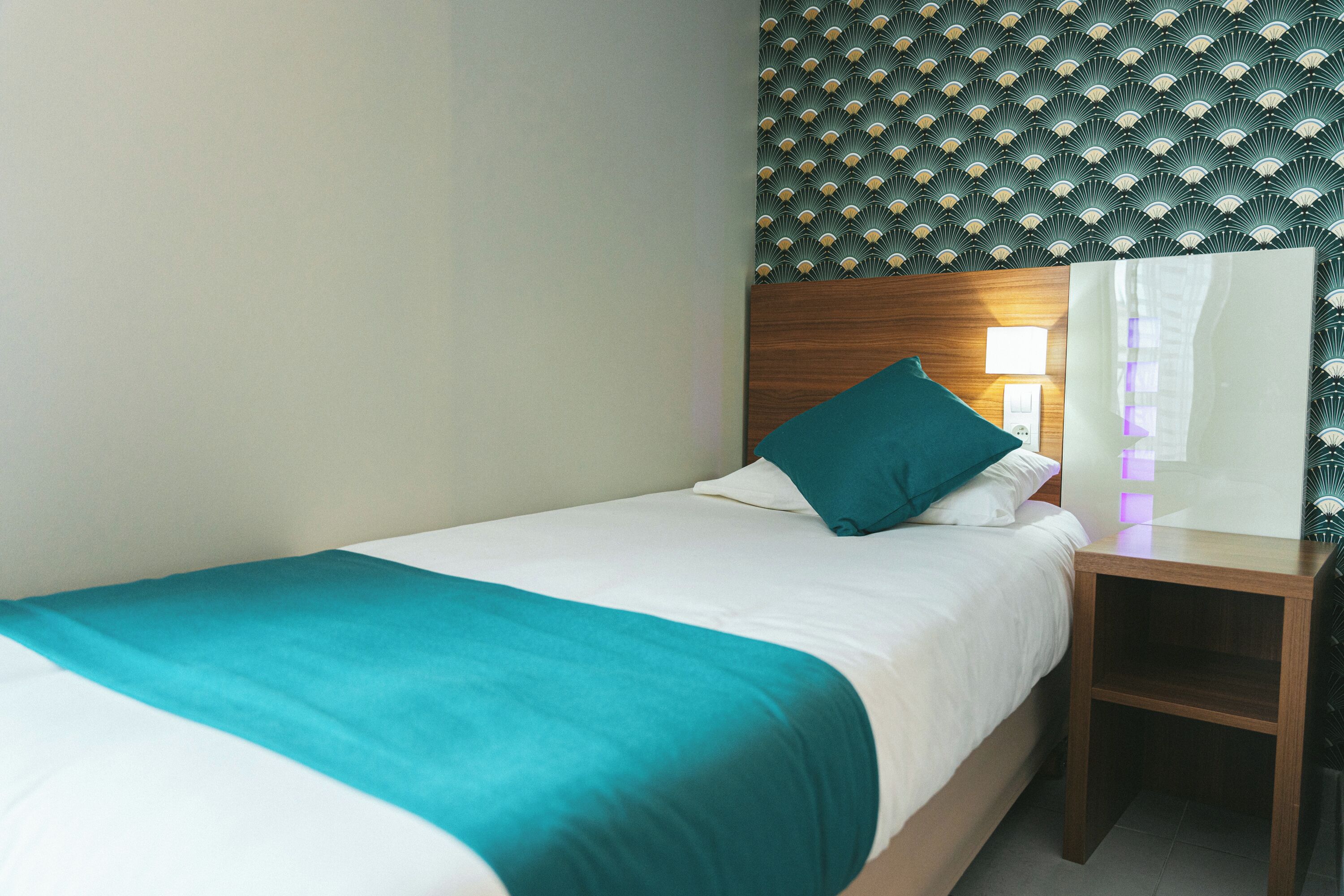A neatly made bed with a teal bedspread and white linens, a wooden nightstand with a lamp, and a patterned wallpaper above the bed.