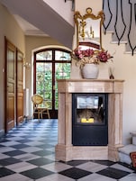 A checkered floor, a fireplace with a vase of flowers, and a chair in a hallway.