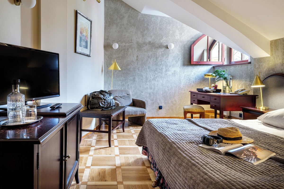A hotel room with a flat-screen TV, a desk with a lamp, a chair, a bed with a blanket, and a bookshelf.