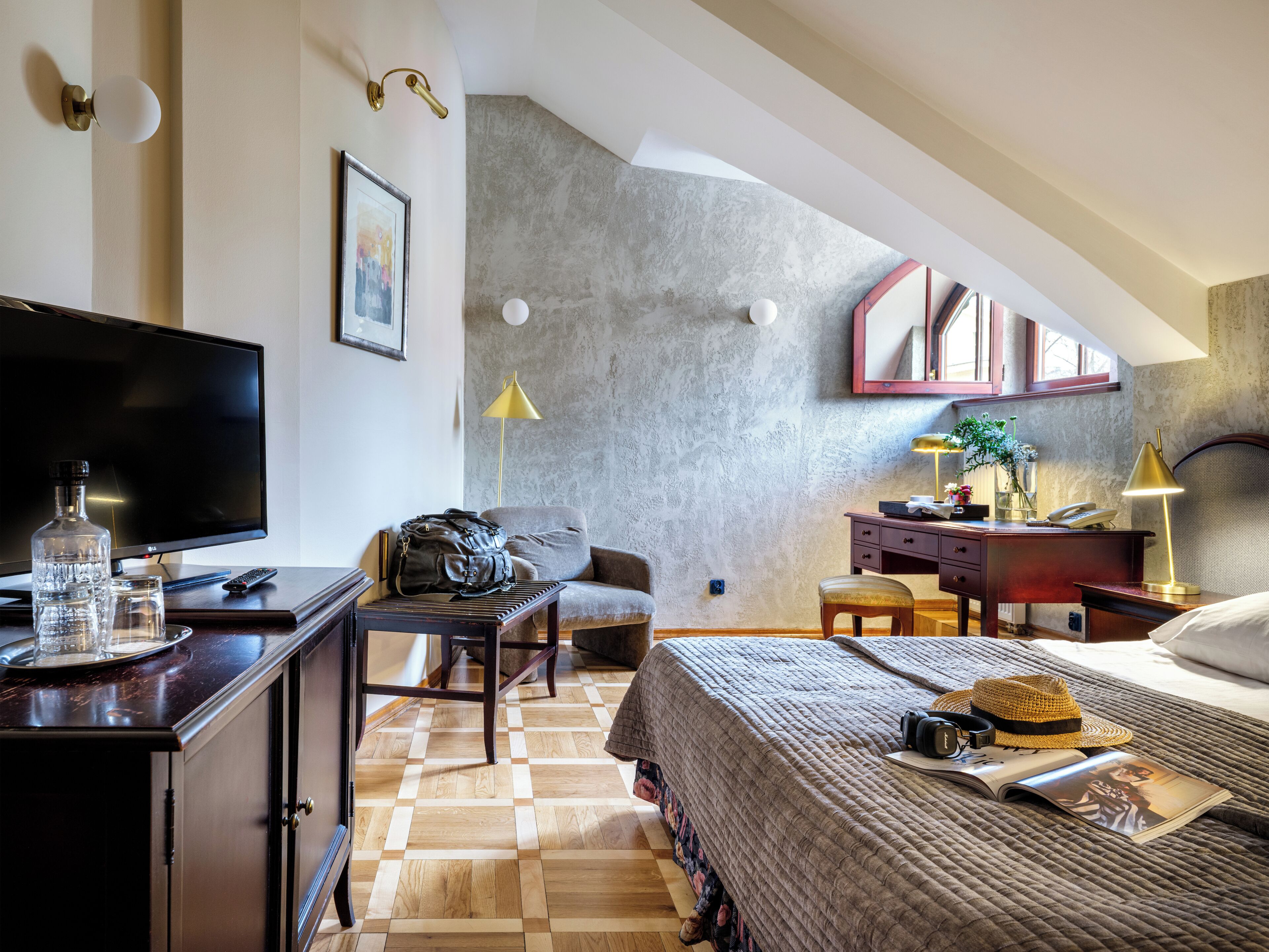 A hotel room with a flat-screen TV, a desk with a lamp, a chair, a bed with a blanket, and a bookshelf.