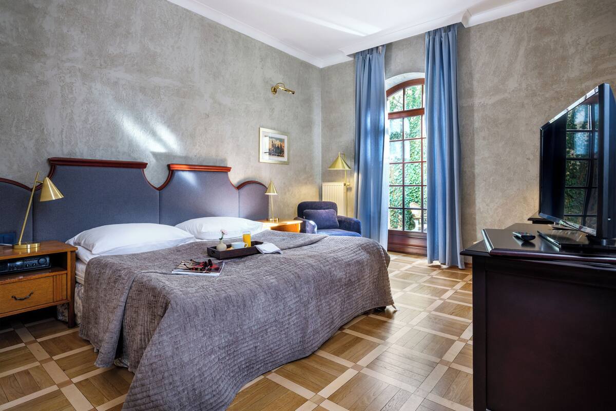 Een hotelkamer met een groot bed, een bureau met een televisie, een stoel, een lamp en een raam met gordijnen.