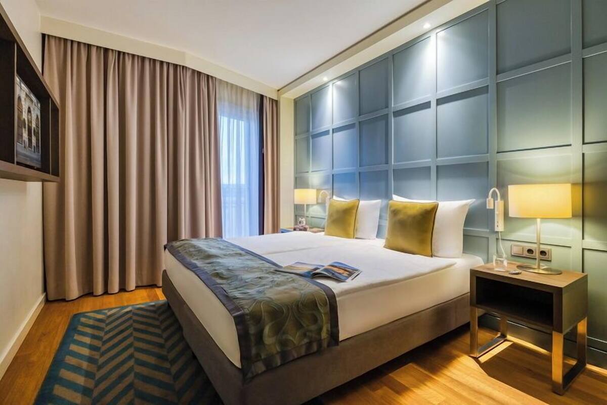 Ein Hotelzimmer mit einem großen Bett, einem Nachttisch, einer Lampe und Blick auf die Stadt durch das Fenster.