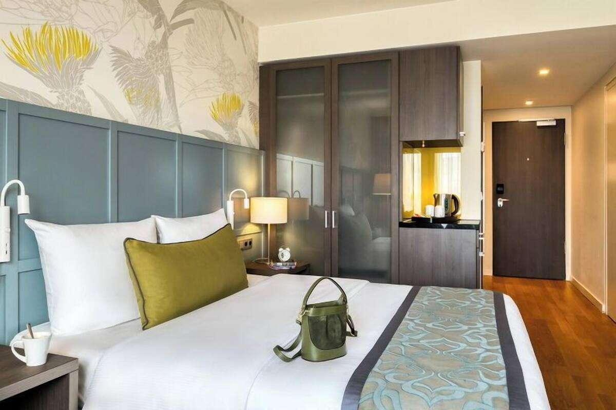 Ein Hotelzimmer mit Bett, Nachttisch, Lampe, Kleiderschrank und einem durch eine Glastür einsehbaren Badezimmer.
