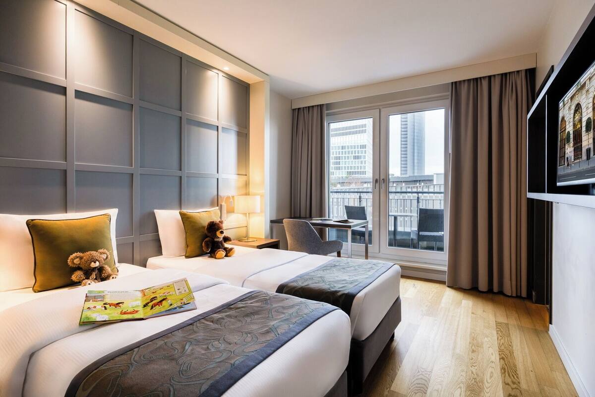 Ein Hotelzimmer mit zwei Betten, einem Schreibtisch, einem Stuhl, einem Fenster mit Blick auf Gebäude und einem an der Wand befestigten Fernseher.