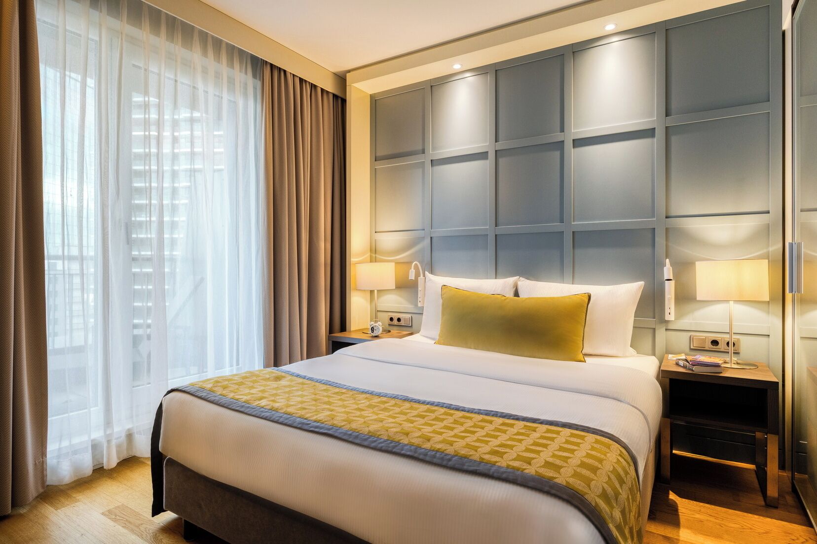 Habitación de hotel con una cama grande, dos mesitas de noche, cabecera y ventana con cortinas.