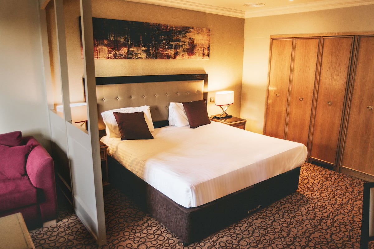 Een hotelkamer met een groot bed, een houten garderobe en een rode fauteuil.