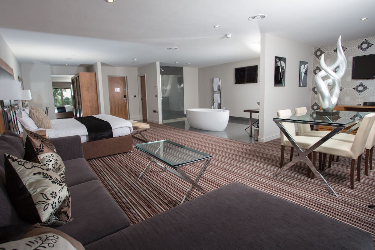 Een moderne hotelkamer met een groot bed, een glazen salontafel, een bank en een zithoek met stoelen.