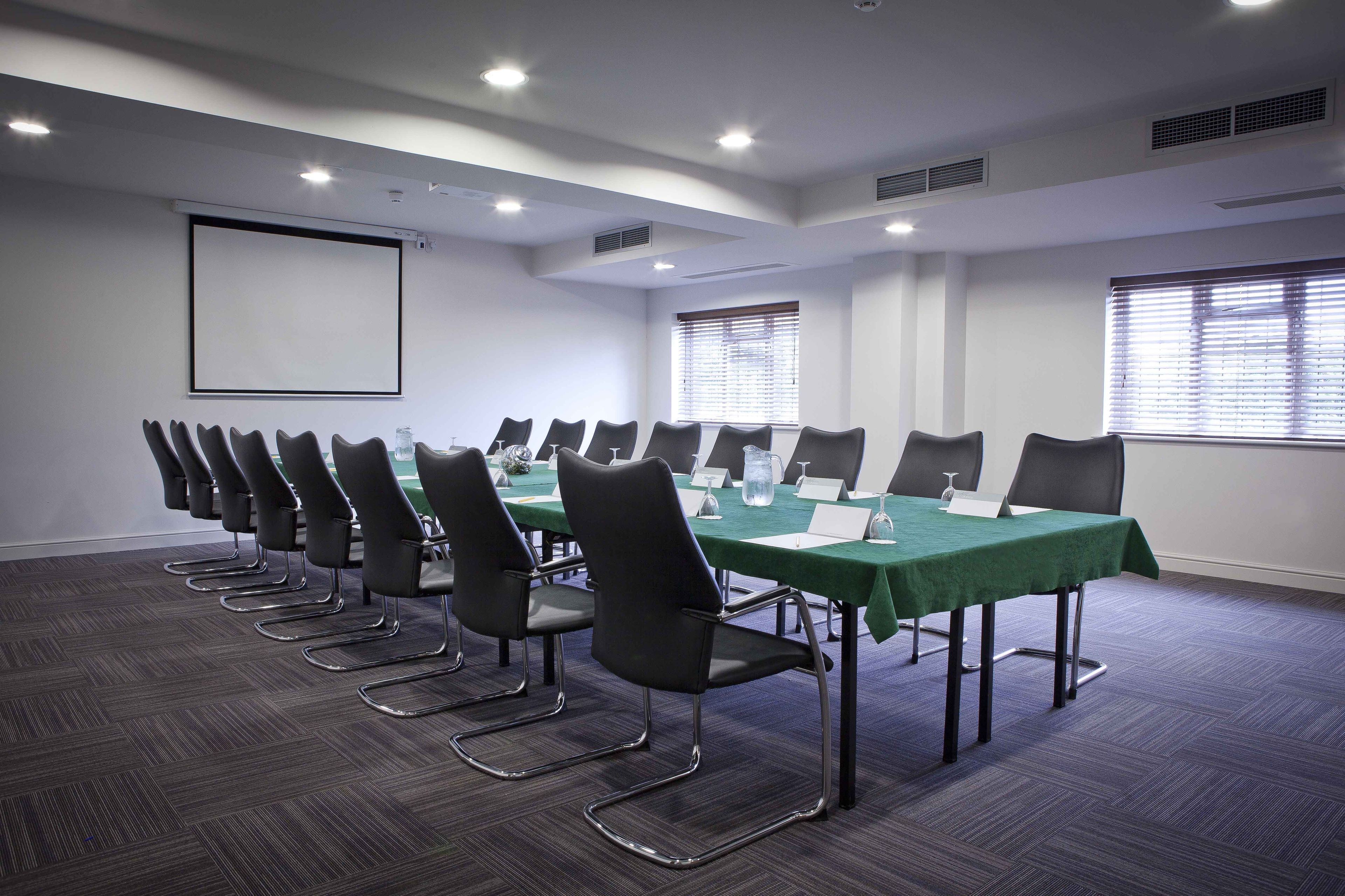 A conference room with a long table, multiple chairs, a projector screen, and windows with blinds.