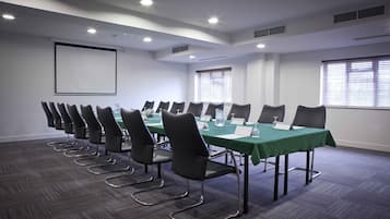 A conference room with a long table, multiple chairs, a projector screen, and windows with blinds.