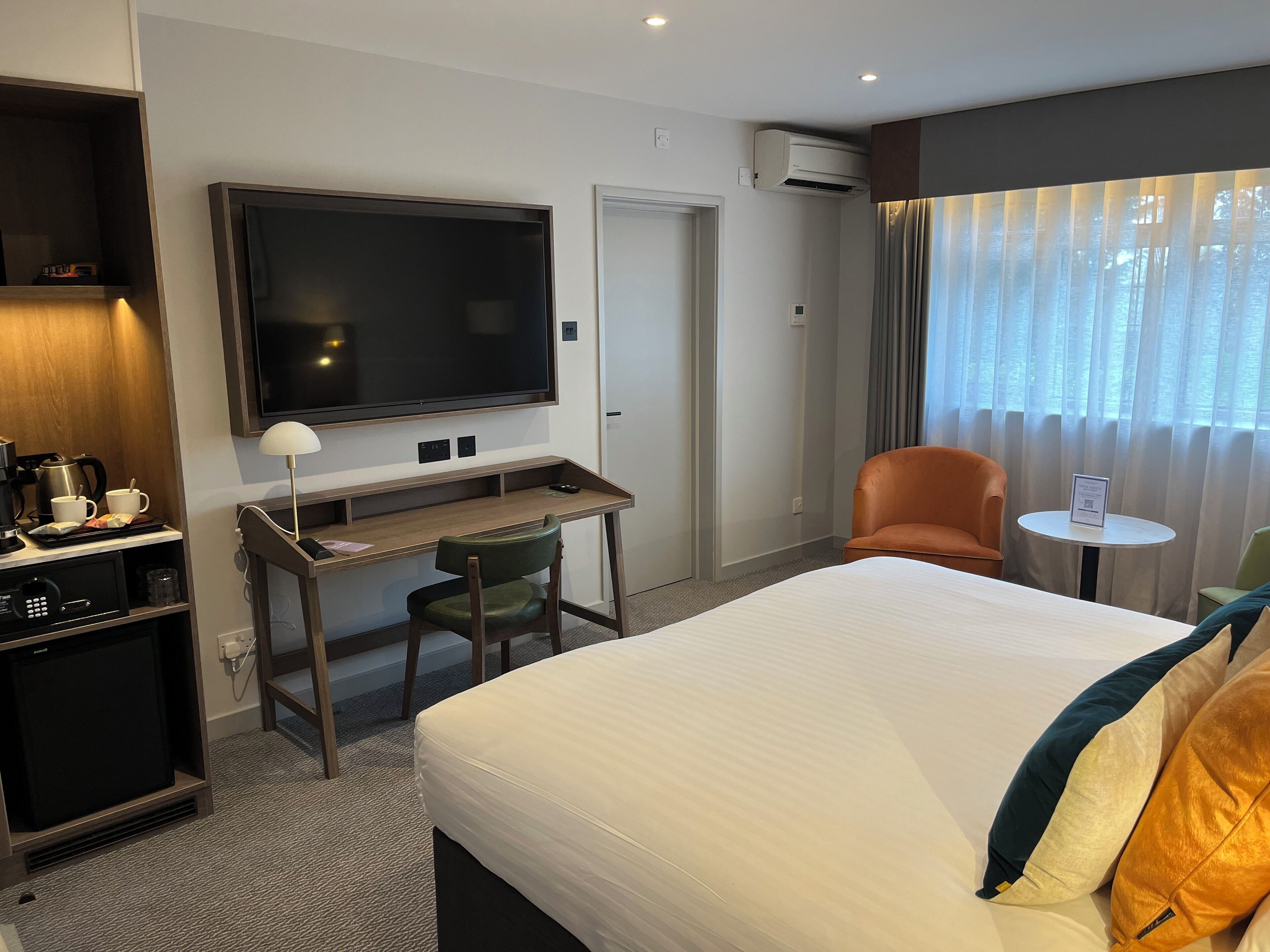 A hotel room with a bed, a desk with a chair, a television, a coffee station, and a window with curtains.