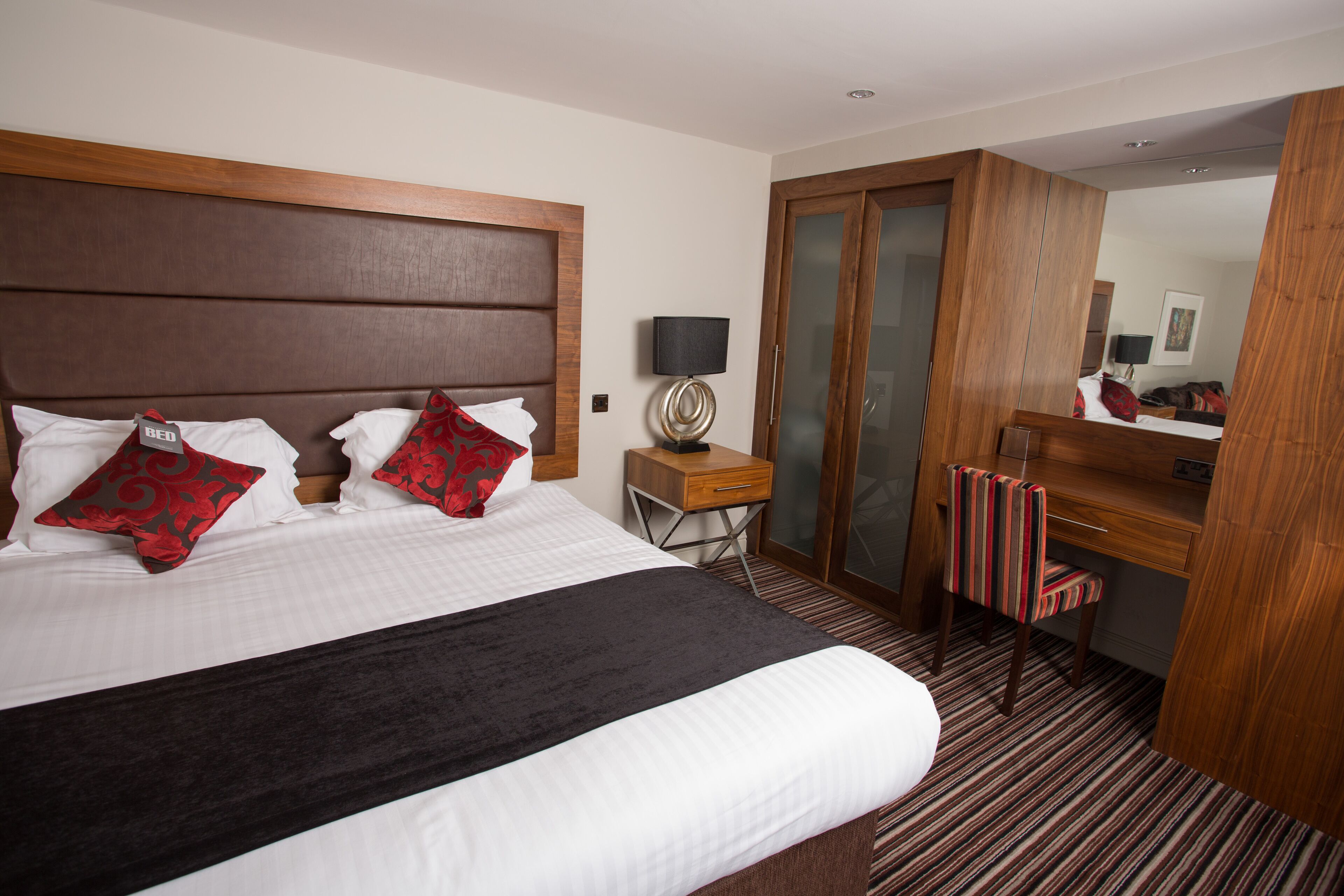 A hotel room with a large bed, a wooden headboard, a desk with a lamp, a mirror, and a striped chair.