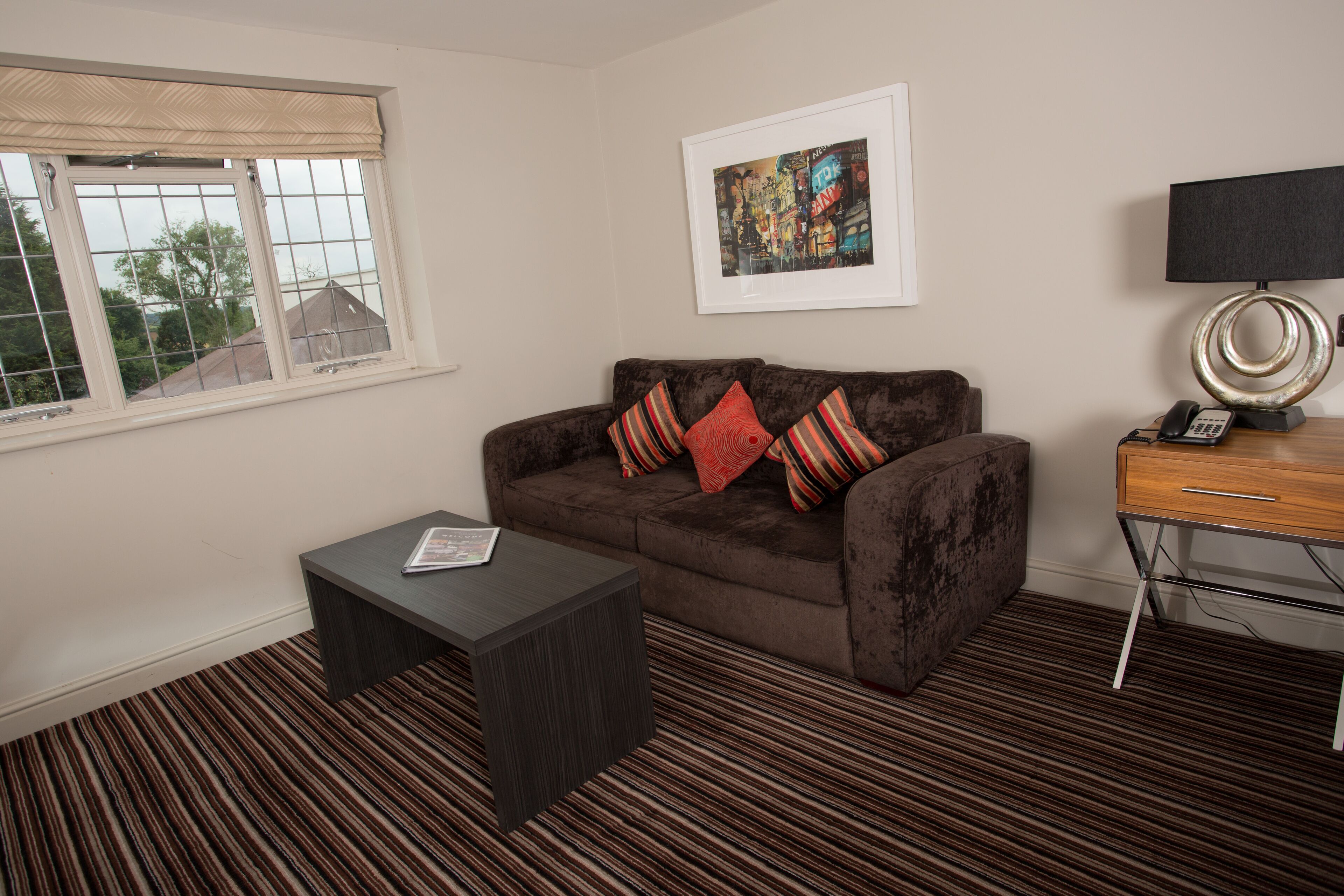 A living room with a brown sofa, a small dark coffee table, a framed picture on the wall, a lamp, and a window with a view of houses.