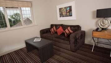 A living room with a brown sofa, a small dark coffee table, a framed picture on the wall, a lamp, and a window with a view of houses.