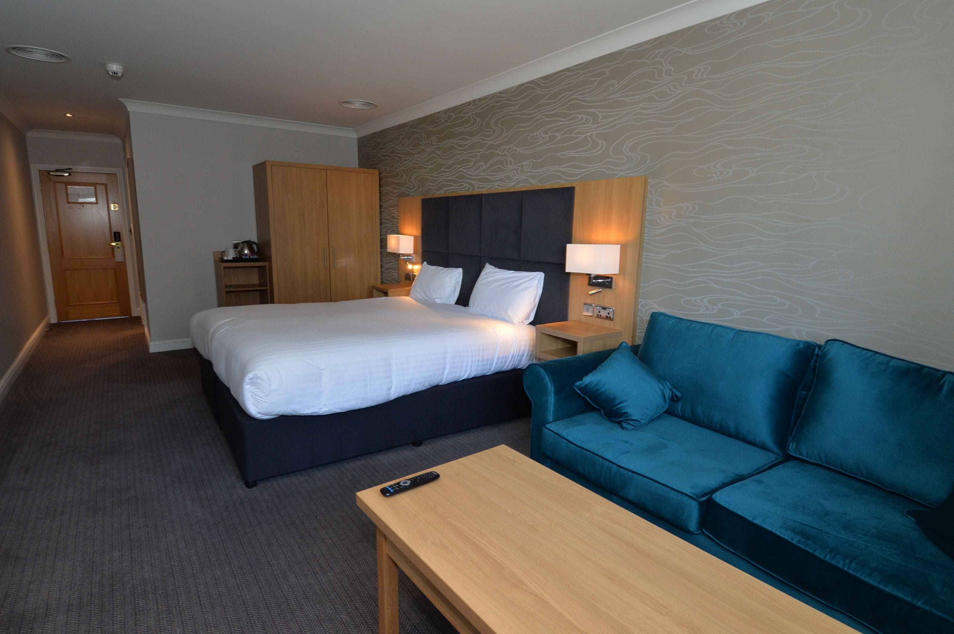A hotel room with a bed, a blue sofa, a wooden table, and a nightstand with a lamp.