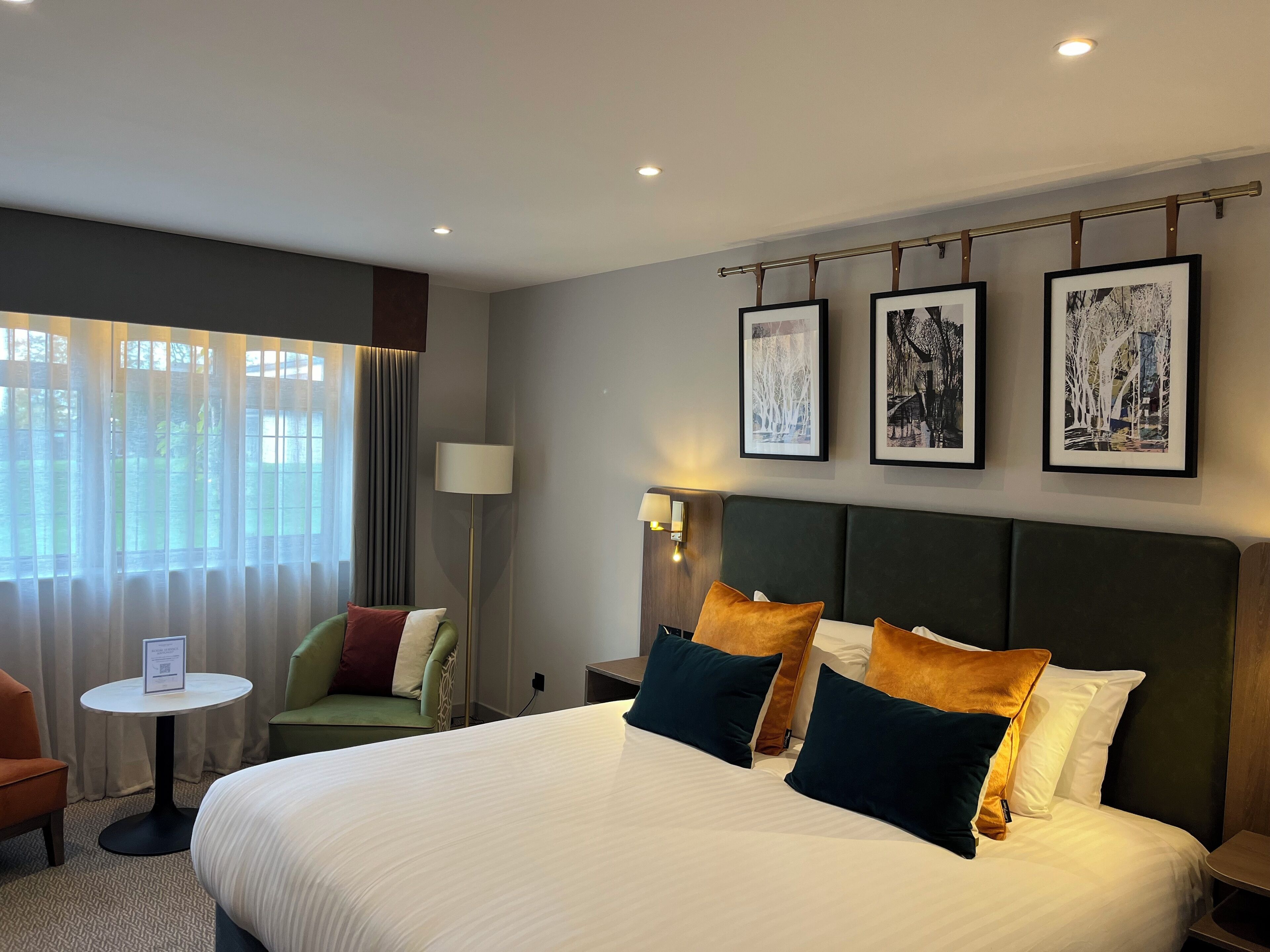 A hotel room with a large bed, a small round table, a green armchair, a floor lamp, and three framed pictures on the wall.