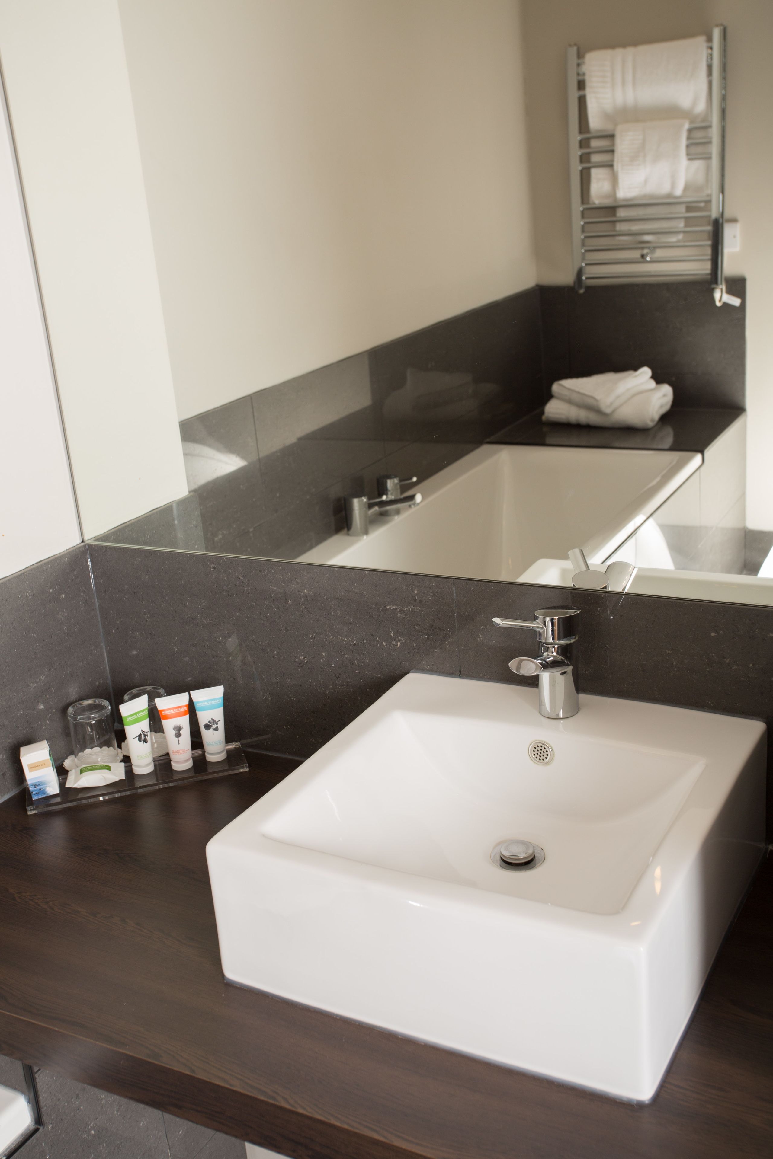 Un baño con un lavamanos blanco, encimera oscura y un espejo que refleja un perchero.