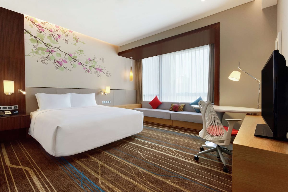 Una habitación de hotel con una cama grande, un escritorio con computadora, televisor, sofá con cojines y una ventana con cortinas.