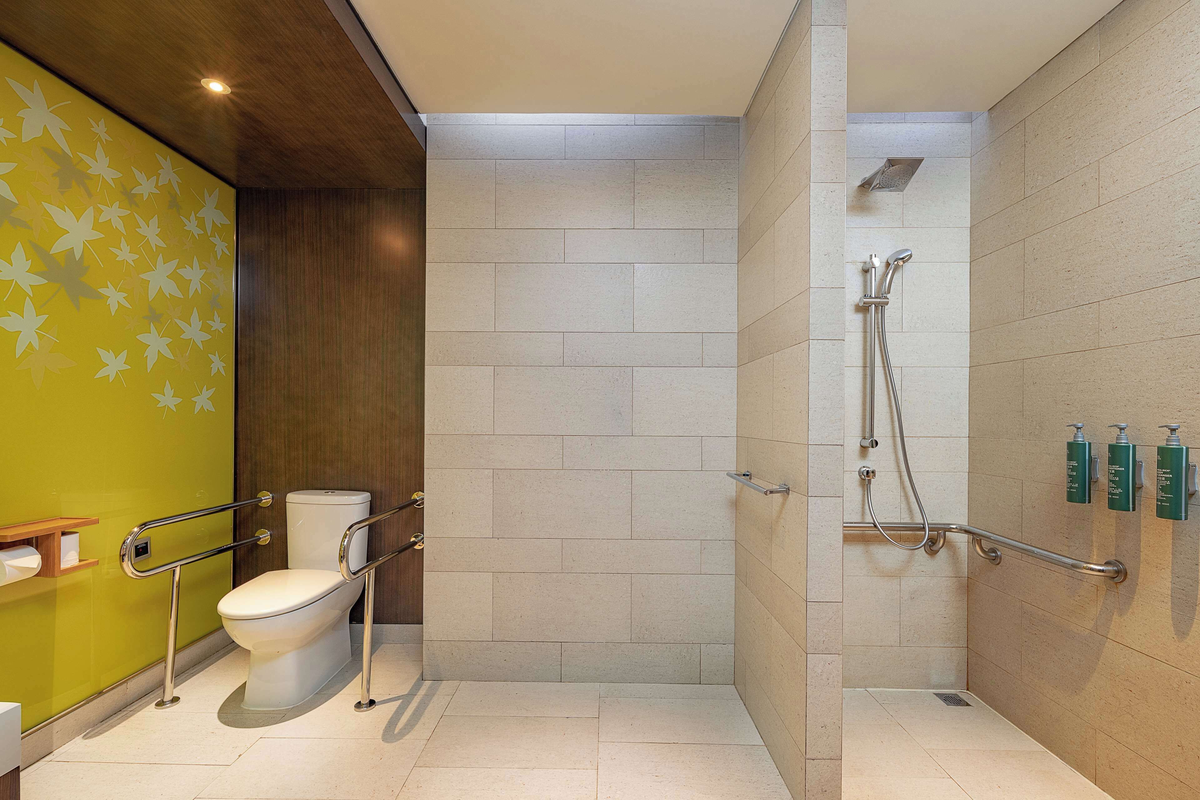 A bathroom with a toilet, a shower with grab bars, and a green wall with leaf patterns.