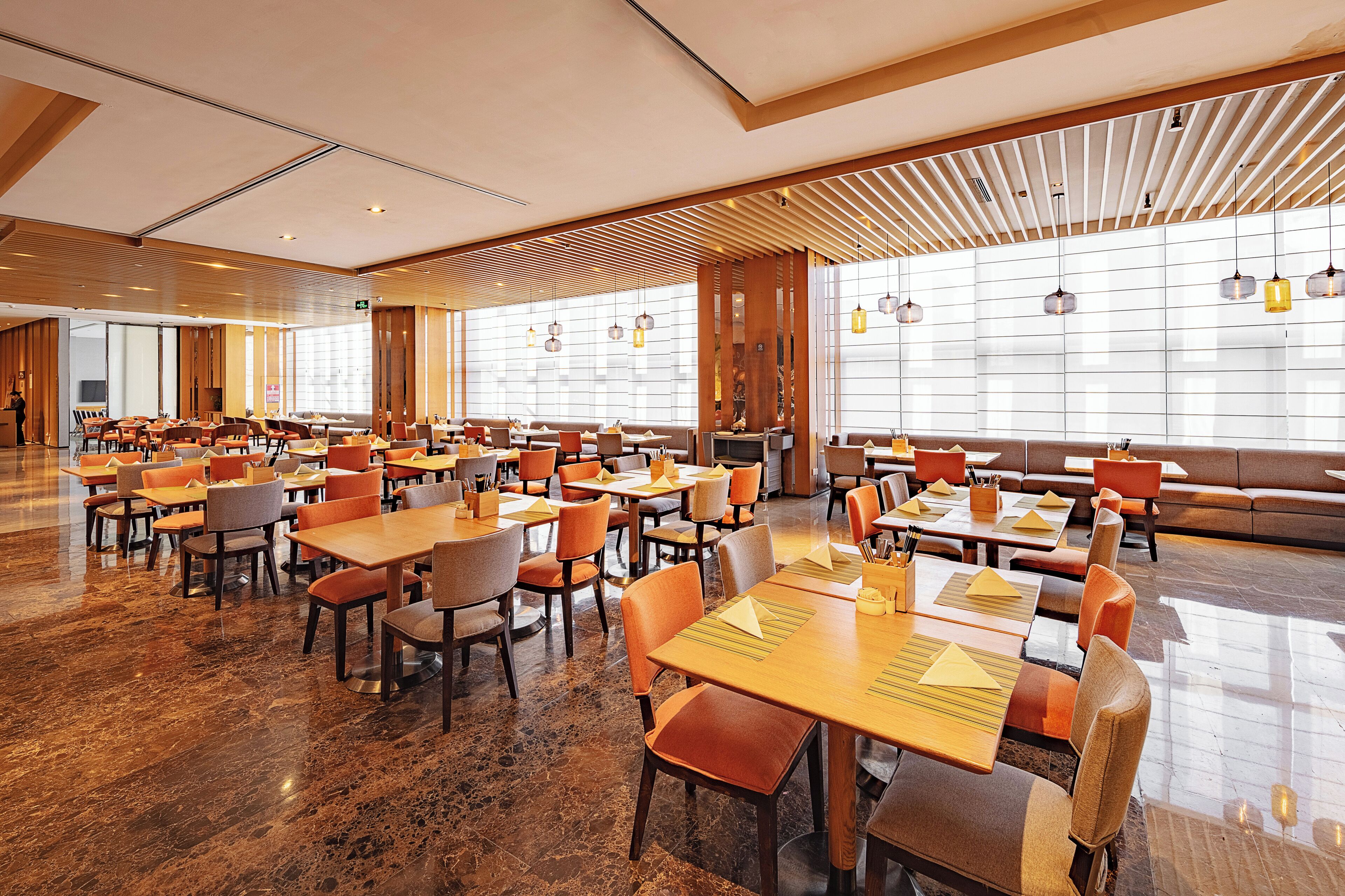 A spacious hotel dining area with multiple tables and chairs, large windows, and a modern interior design.