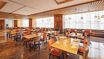 A spacious hotel dining area with multiple tables and chairs, large windows, and a modern interior design.