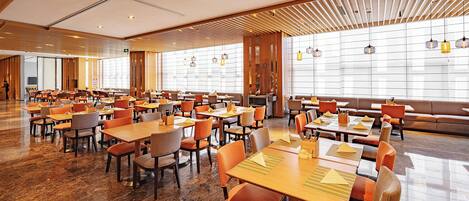 A spacious hotel dining area with multiple tables and chairs, large windows, and a modern interior design.